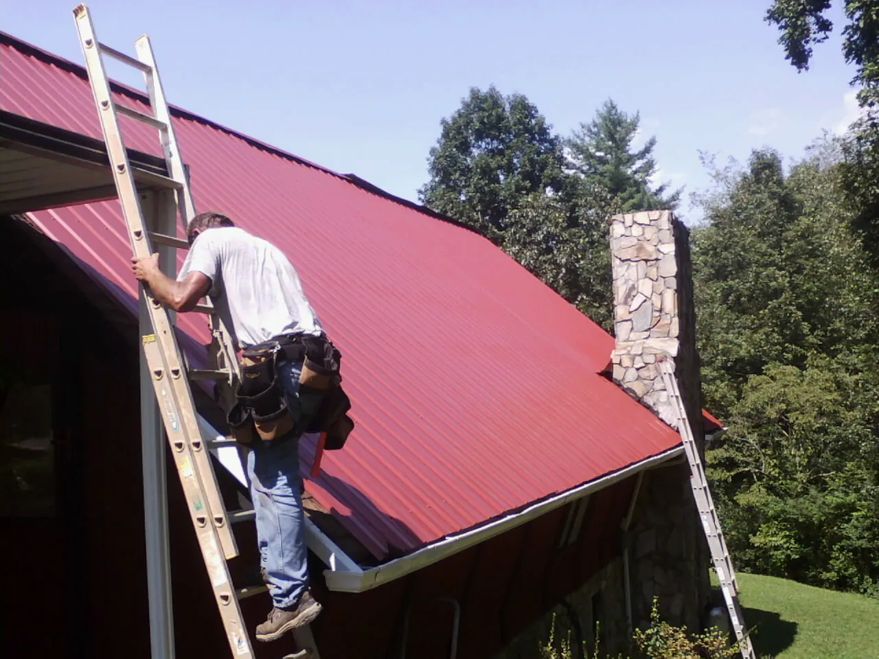 Roofer on a ladder beside a red metal roof and stone chimney — Soffit & Fascia Repair by Roofing Brothers in Proctor